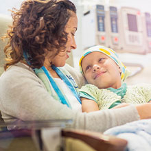 Woman sits holding young sick girl on her lap in hospital room.